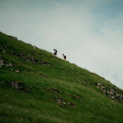 Two walkers climbing a steep green hillside under a pale sky