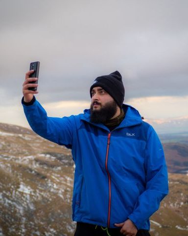 A bearded man in a blue waterproof jacket and woollen hat taking a selfie on a winter hillside