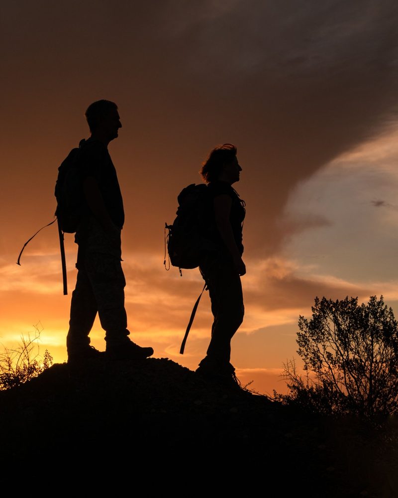 Two walkers with rucksacks silhouetted against an orange sunset on a hilltop