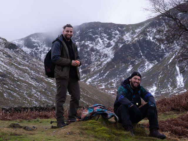 Two men laughing on a snowy hillside, one standing with a rucksack and one sitting on the grass