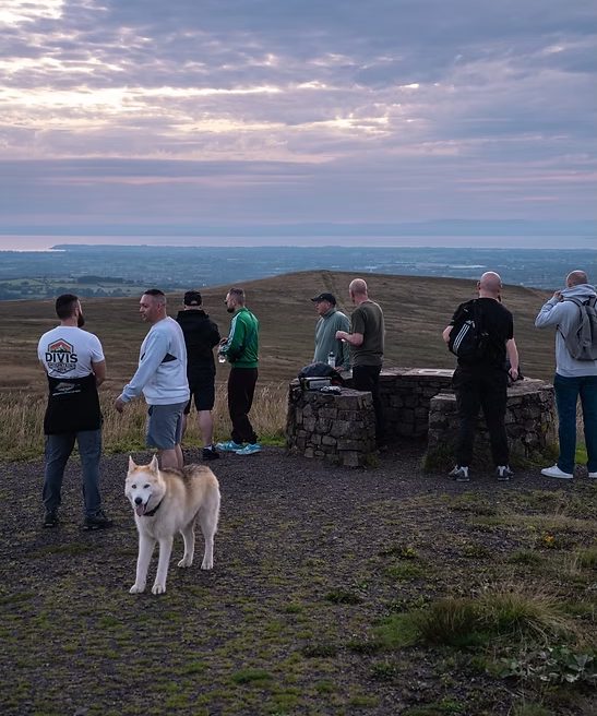 Men gathered at the viewpoint on Divis Mountain at dusk, talking in small groups, with a husky in the foreground